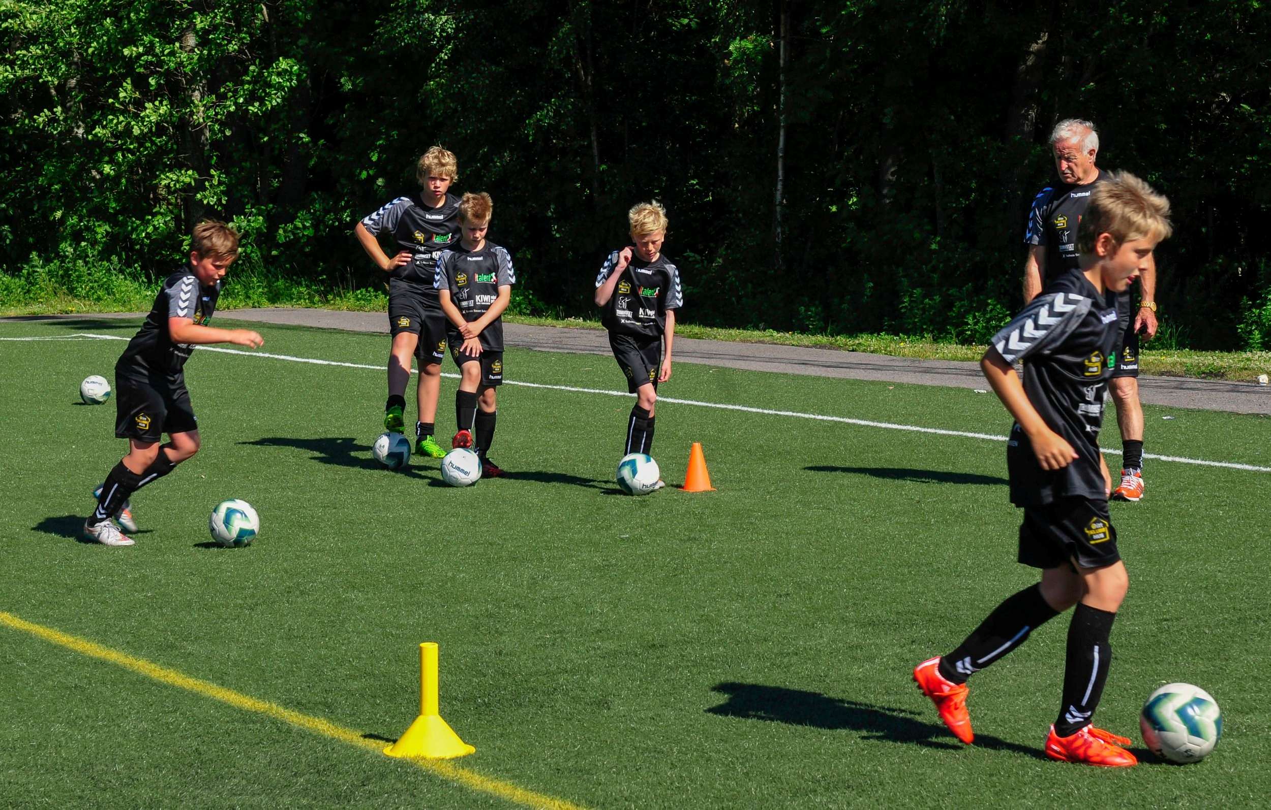 Unge spillere i sort Italent-drakt fører ball mellom kjegler på gressbanen
